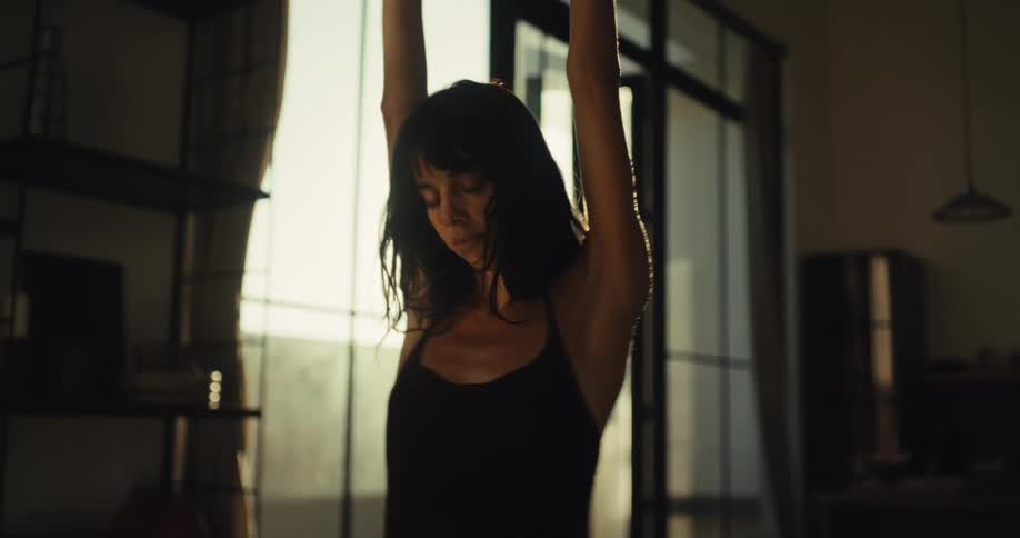 A beautiful woman dances freely and expressively in slow motion in her dimly lit apartment, backlit by the warm, golden light of the sun