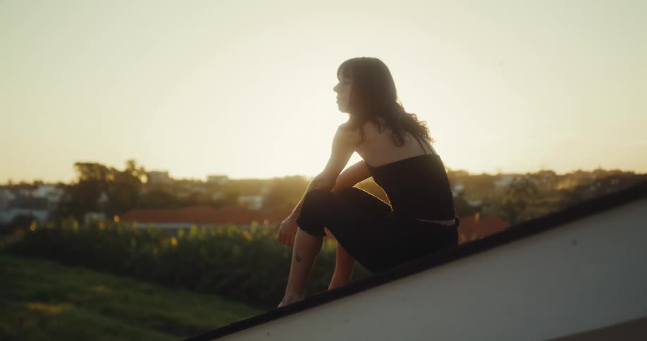 A lone woman sits on a rooftop, silhouetted against the warm glow of the setting sun