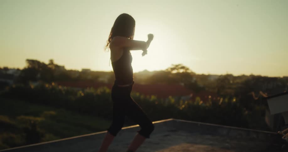 Slow motion view of a woman dancing barefoot on a rooftop, silhouetted against the beautiful golden hour sunset