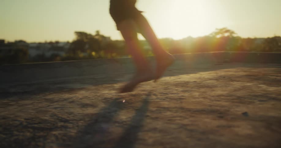 A low-angle, slow-motion shot of a person's bare feet jumping rope on a rooftop at sunset