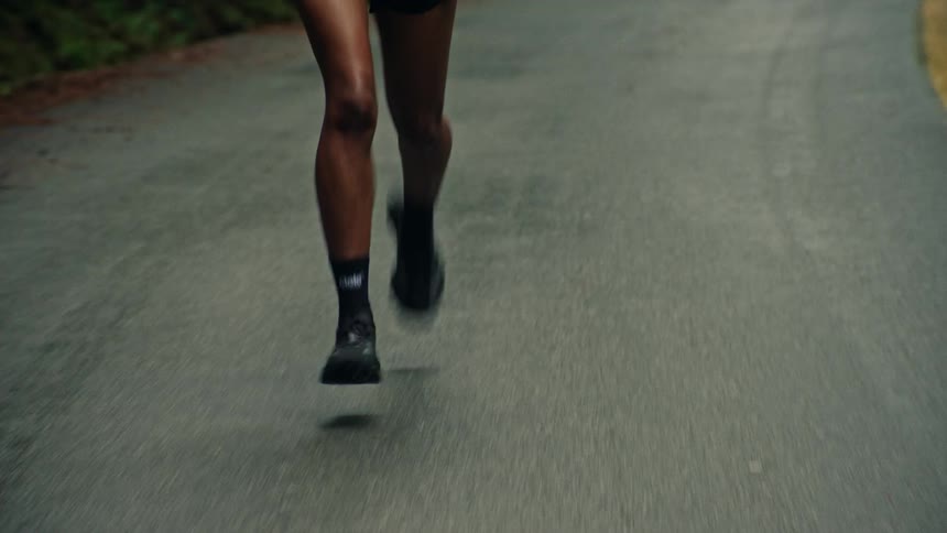 A close-up front view of an athletic woman's legs running on a wet asphalt road surrounded by lush green nature