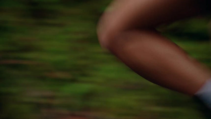 A close-up side-view tracking shot follows a fit woman's torso and legs as she runs at speed along a road in a lush green forest