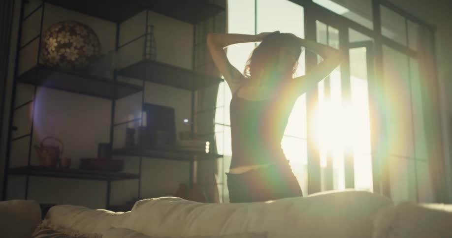 A woman in pajamas stretches and dances slowly in her sun-drenched living room in the morning
