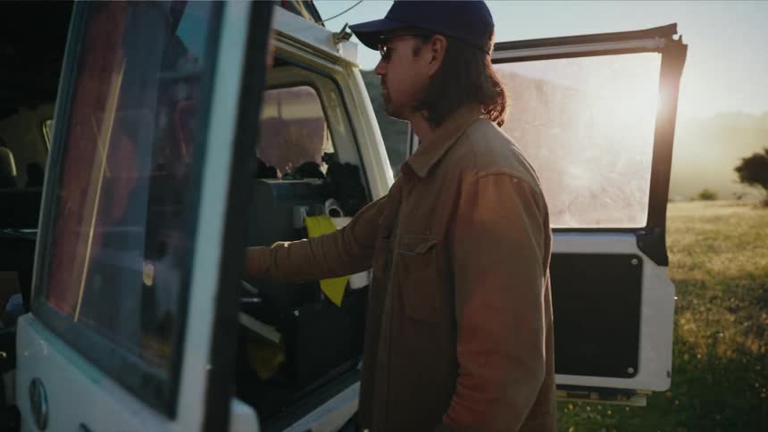 A man unloads a folding table from his vintage camper van to set up camp in a scenic meadow at sunset