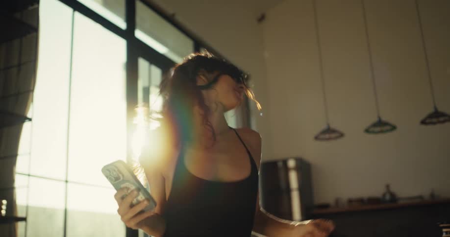 A joyful woman dances energetically around her sun-drenched apartment while looking at her phone