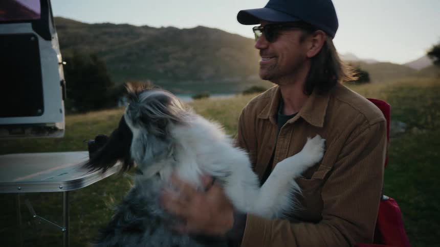 A happy man sits on a camp chair and affectionately pets his Australian Shepherd on his lap while on a van life trip at sunset