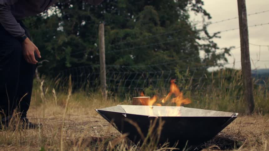 Slow motion shot of a man using a hatchet to adjust a log in a portable campfire in a grassy field