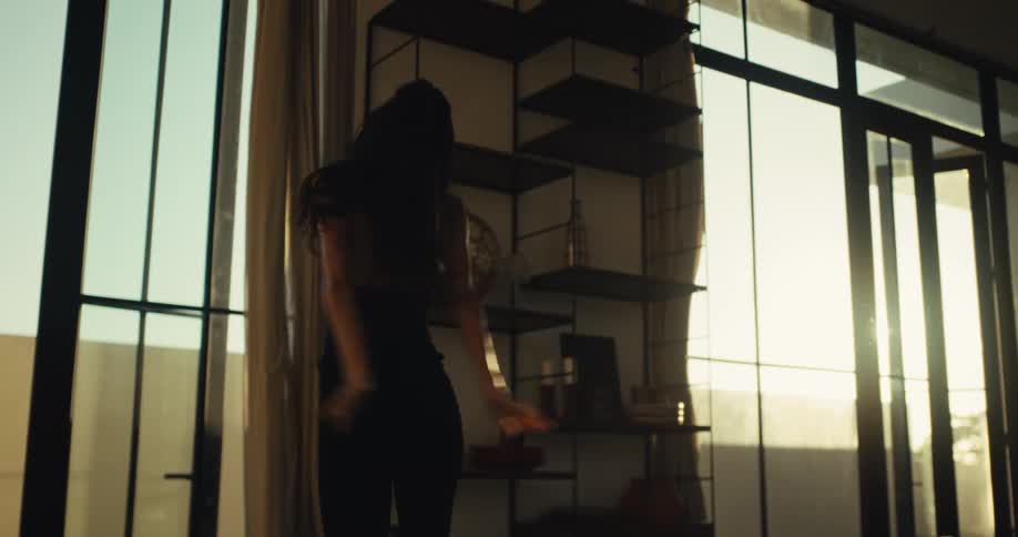 A woman in a black outfit dances energetically with pure joy on a sofa in her sunlit modern apartment before falling back to rest