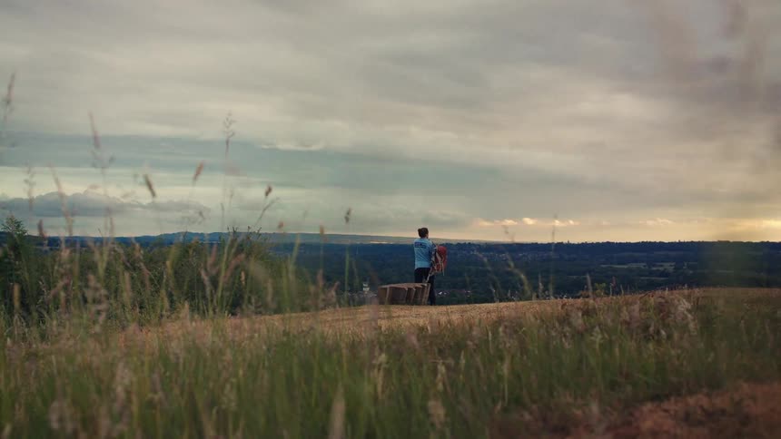 A lone hiker with a backpack stands on a grassy hilltop at dusk, gazing out at the expansive valley below under a dramatic, cloudy sky