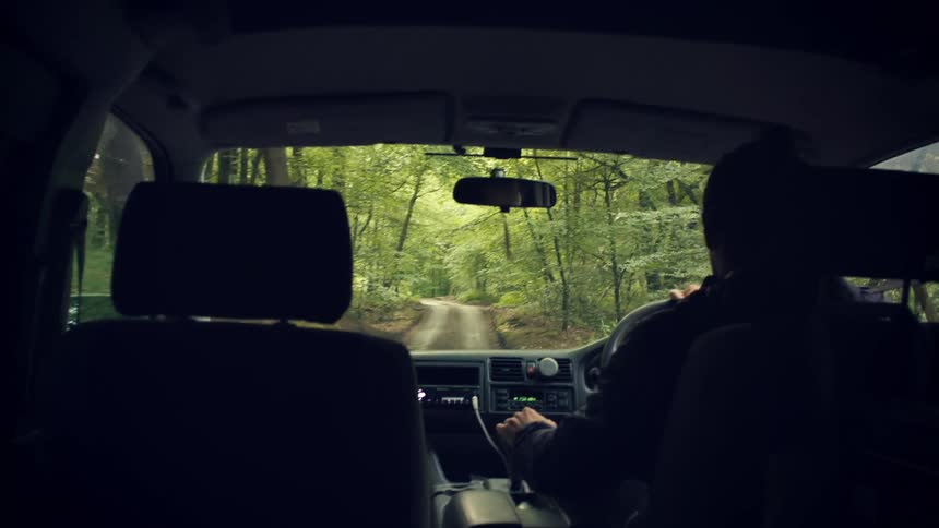 Interior car view of a person driving slowly down a narrow dirt road surrounded by a lush green forest