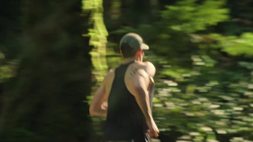 An athletic man with an Olympic tattoo runs with determination on a scenic trail through a sunlit forest
