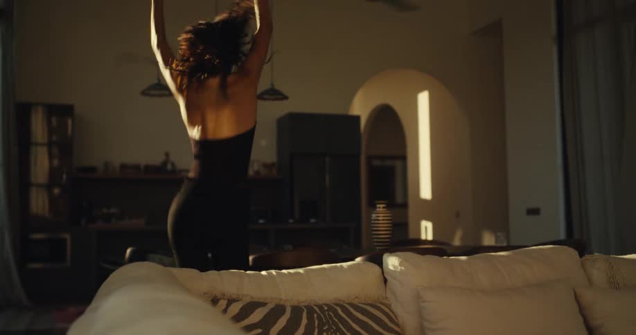 A carefree woman in a black top dances with abandon on her couch in a sunlit room, captured in slow motion