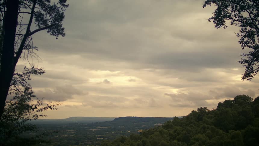 A vast, lush green valley unfolds under a dramatic, overcast sky at dusk, framed by dark trees