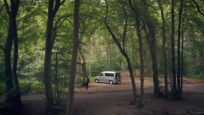 A person with a backpack walks towards a silver camper van parked in a clearing in a dense green forest