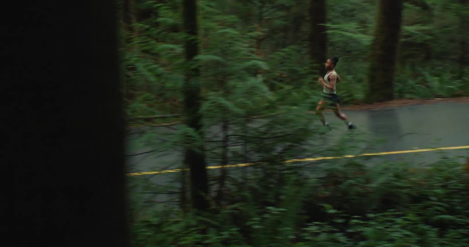 A tracking shot follows a woman running on a secluded road that winds through a dense, green forest
