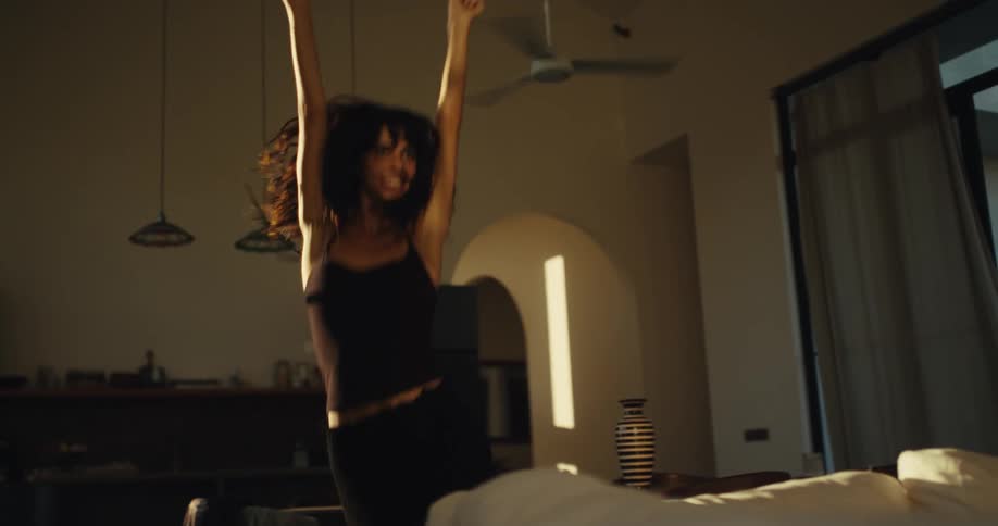 A carefree young woman with dark hair joyfully dances in slow motion in her sunlit living room