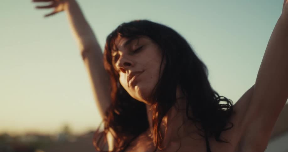 A beautiful young woman with her eyes closed listens to music on wireless earbuds, stretching and enjoying the golden hour sunlight