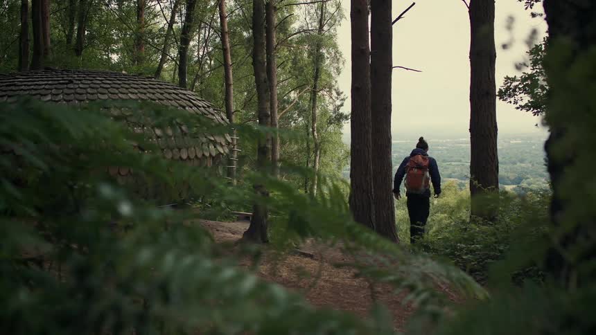 A hiker with a backpack stands at the edge of a forest next to a wooden yurt, looking out at the expansive view below