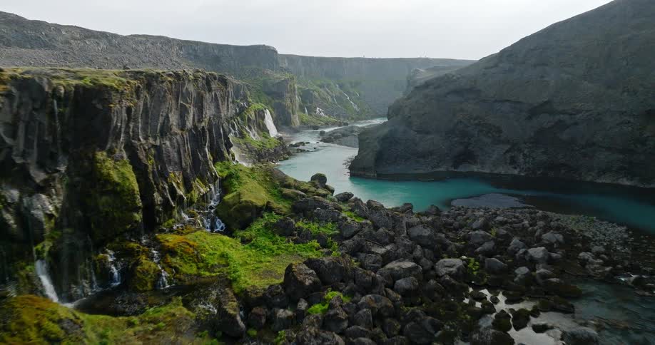 Cinematic aerial footage flies slowly down into a majestic, rocky canyon revealing numerous waterfalls and a striking turquoise glacial river