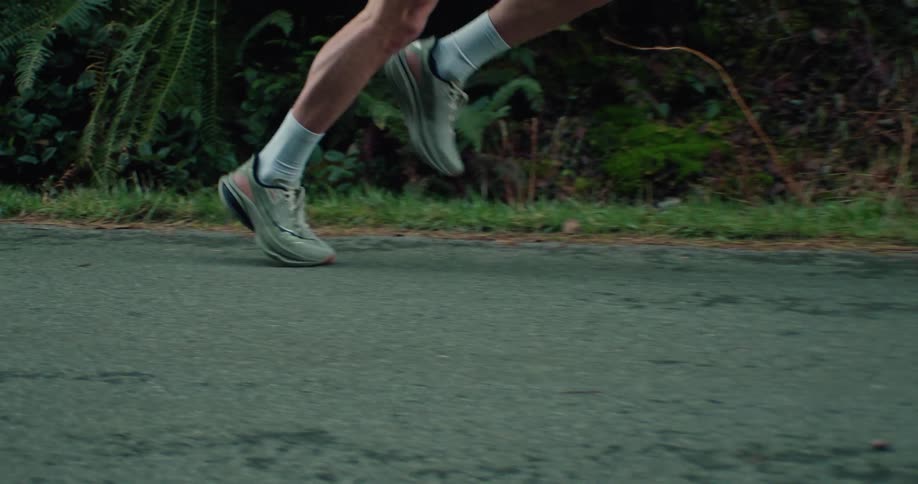 The powerful stride of a runner is captured in extreme slow motion, focusing on their muscular legs and feet on a paved road
