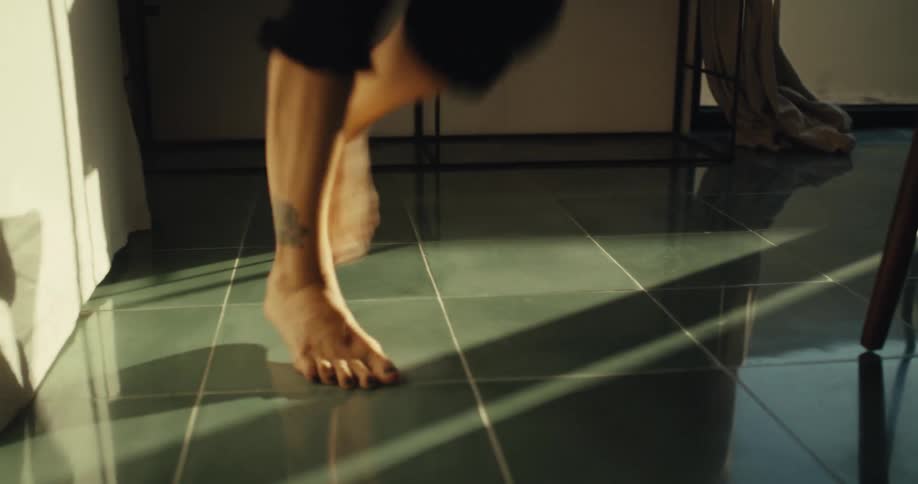 Close-up of a tattooed person's bare feet dancing gracefully on a reflective tiled floor in the warm sunlight