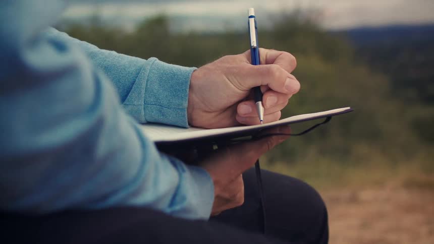 Close-up of a person's hand writing with a mechanical pencil in a journal while sitting outdoors in a natural setting