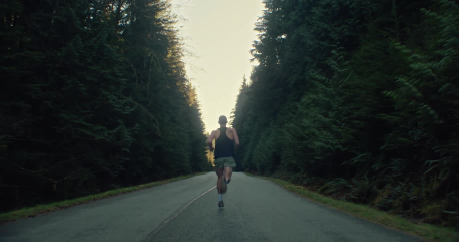 A determined male athlete runs down the middle of a remote road surrounded by a dense evergreen forest