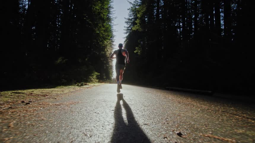 A cinematic slow-motion shot follows a determined male athlete as he runs down a paved forest road into the bright morning sun