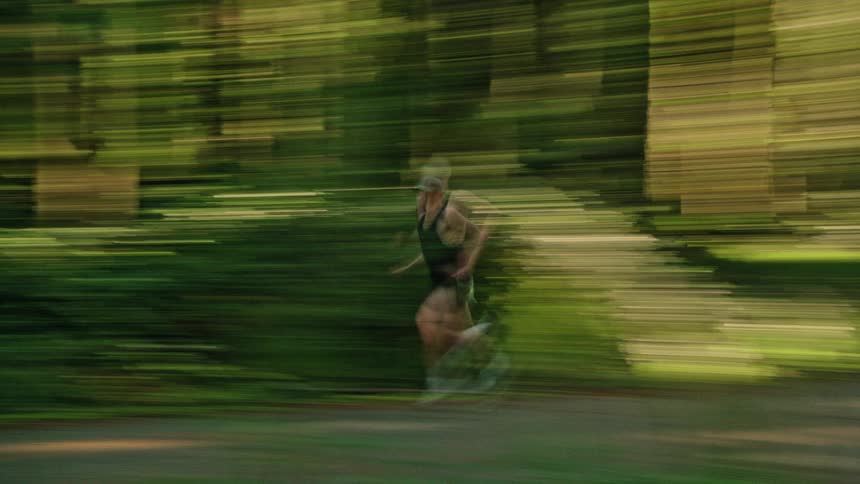 An artistic motion blur shot follows a man as he runs on a paved trail through a lush green forest