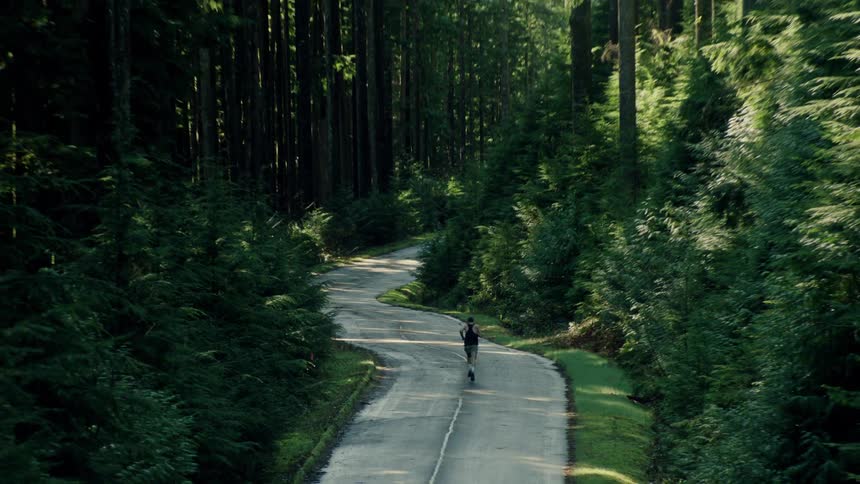A high angle shot follows a man running down a winding road, pulling back to reveal the vast, dense green forest surrounding him