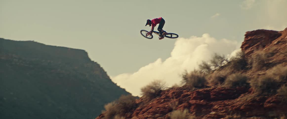 A professional mountain biker launches off a red rock cliff in slow motion, soaring through the air before landing on the steep descent