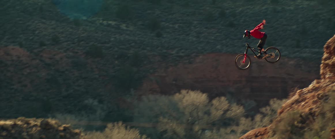 An extreme mountain biker launches off a rocky desert cliff, performing a trick in slow motion before landing and speeding down the dusty trail