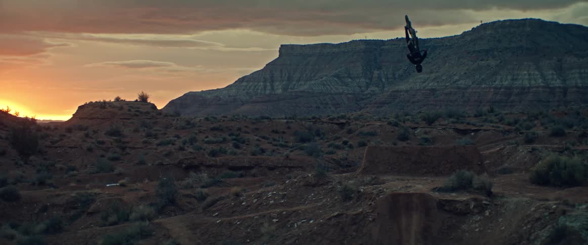 A silhouetted mountain biker performs a dramatic backflip trick in slow motion off a dirt jump with a vast desert mesa landscape in the background at dusk