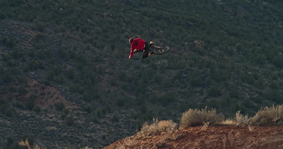 A mountain biker in a red shirt launches off a dirt ramp in the desert, performing a stylish tabletop trick in slow motion