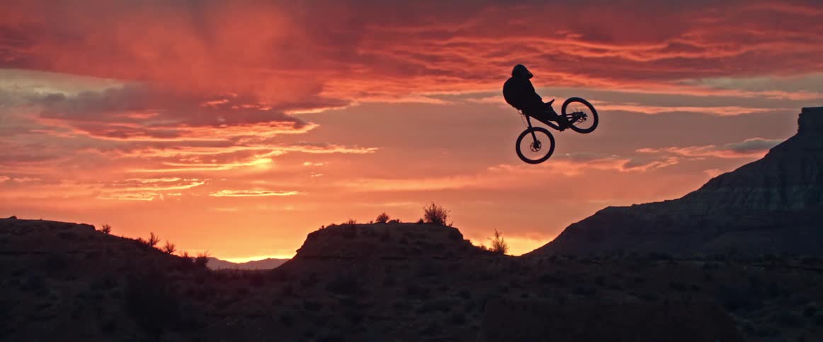 In slow motion, the silhouette of a mountain biker soars through the air off a dirt jump against a stunning desert sunset
