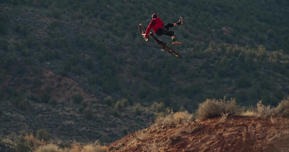 A mountain biker flies through the air in slow motion, performing a no-footed trick off a large dirt jump in a desert landscape