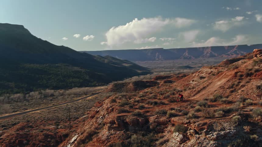 An aerial view follows a mountain biker navigating a thrilling trail along a red rock ridge in a vast desert canyon