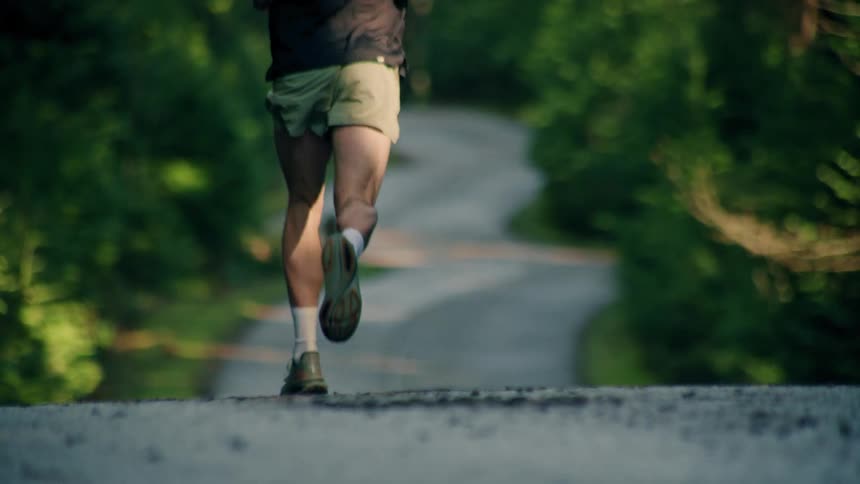 Slow motion rear view of a fit male runner's legs as he jogs down a paved road surrounded by lush green trees