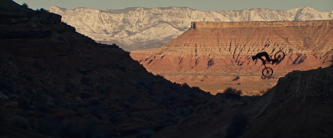 A silhouetted mountain biker soars through the air in slow motion while performing a stunt with a dramatic desert and snow-capped mountain backdrop