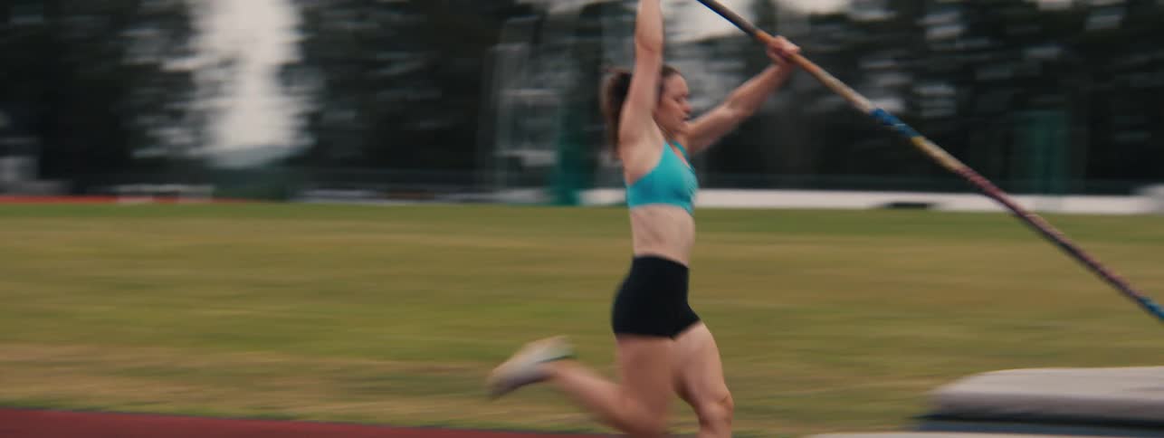 A determined female pole vaulter sprints down an athletics track and successfully clears the bar in a slow-motion action shot