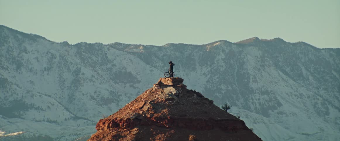 A lone mountain biker pauses on a dramatic red rock peak to admire the stunning vista of snow-covered mountains