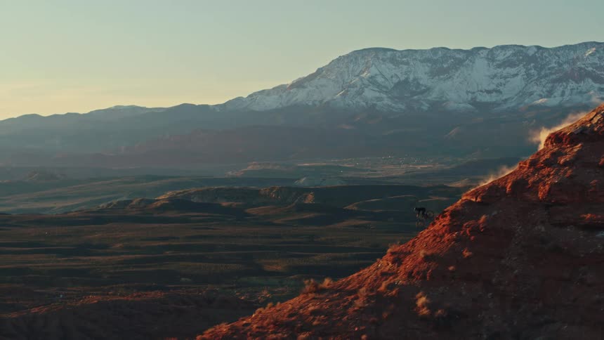 A mountain biker descends a steep, dusty red rock cliff at sunset with a vast desert valley and snow-capped mountains in the background