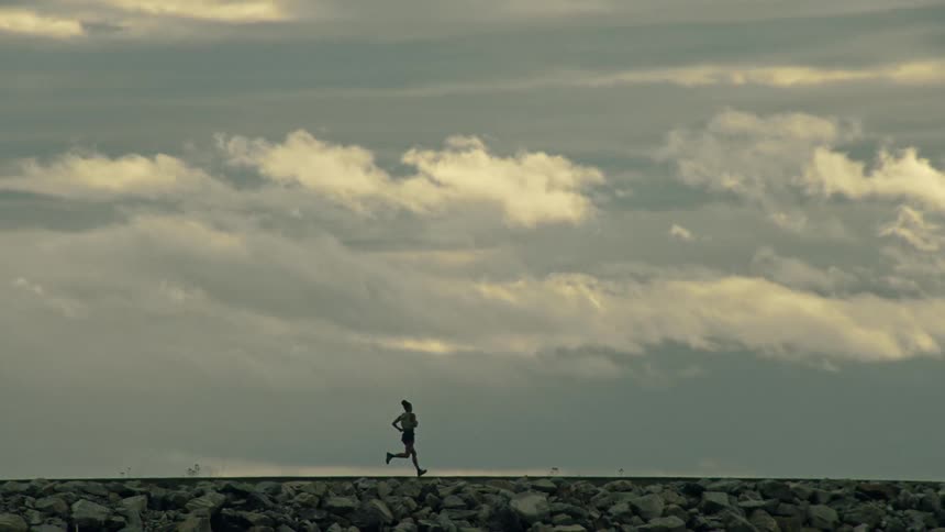 A woman's silhouette is captured in slow motion as she runs along a rocky breakwater under a dramatic, cloudy sky