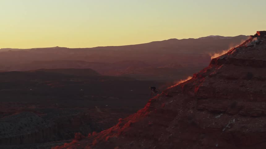 Epic aerial tracking shot of a lone mountain biker descending a narrow, dusty ridge of a red rock butte at sunset