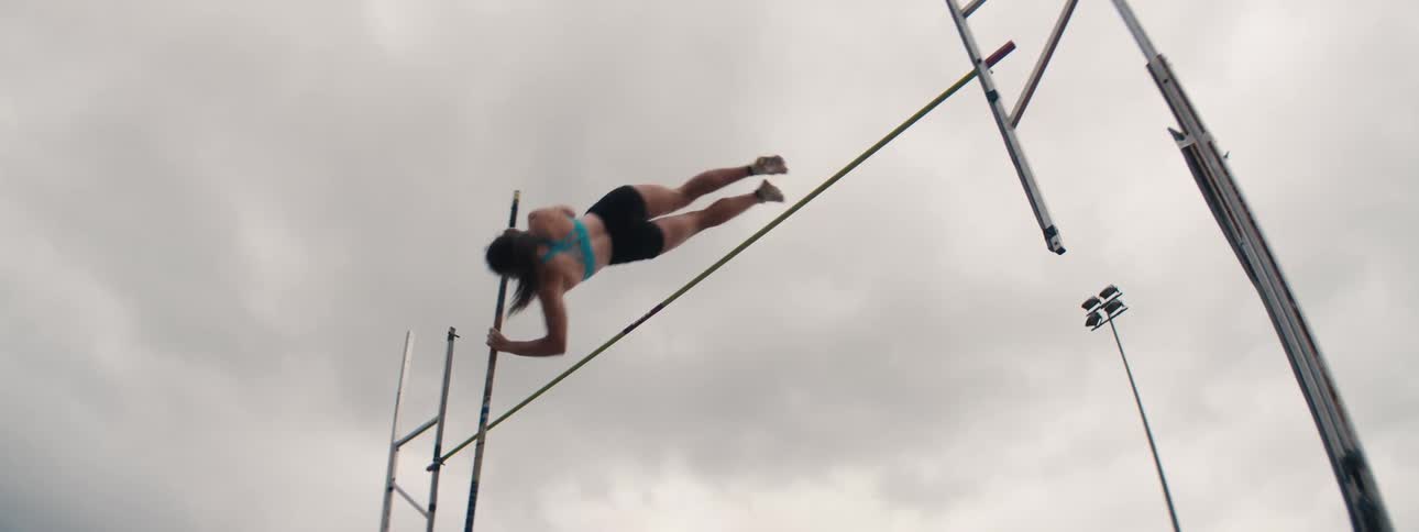 A female athlete clears the bar in a dramatic low-angle, slow-motion pole vault