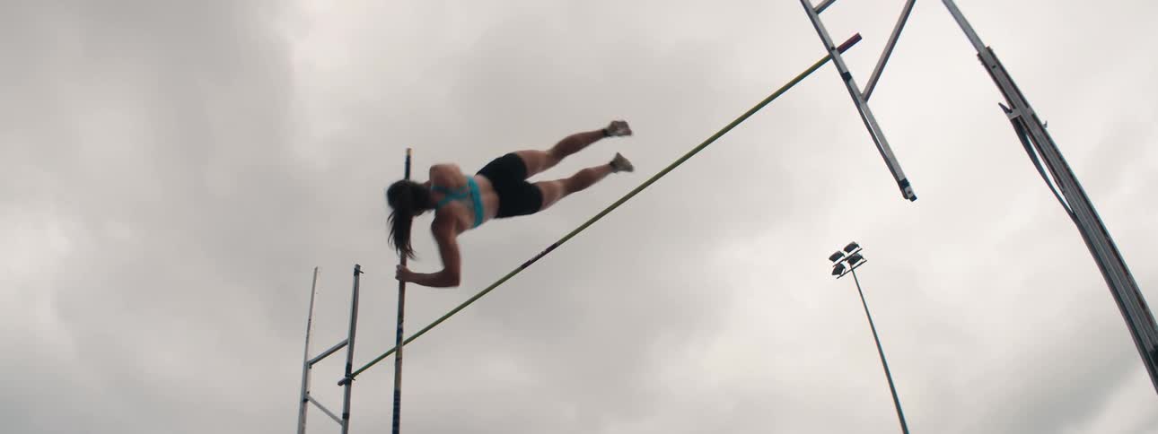A powerful female athlete soars through the air in slow motion as she successfully clears the bar during a pole vault jump