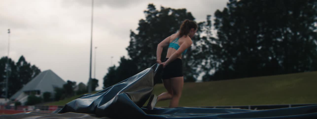 A strong female athlete pulls a heavy protective cover over a landing pad on an athletics track in slow motion