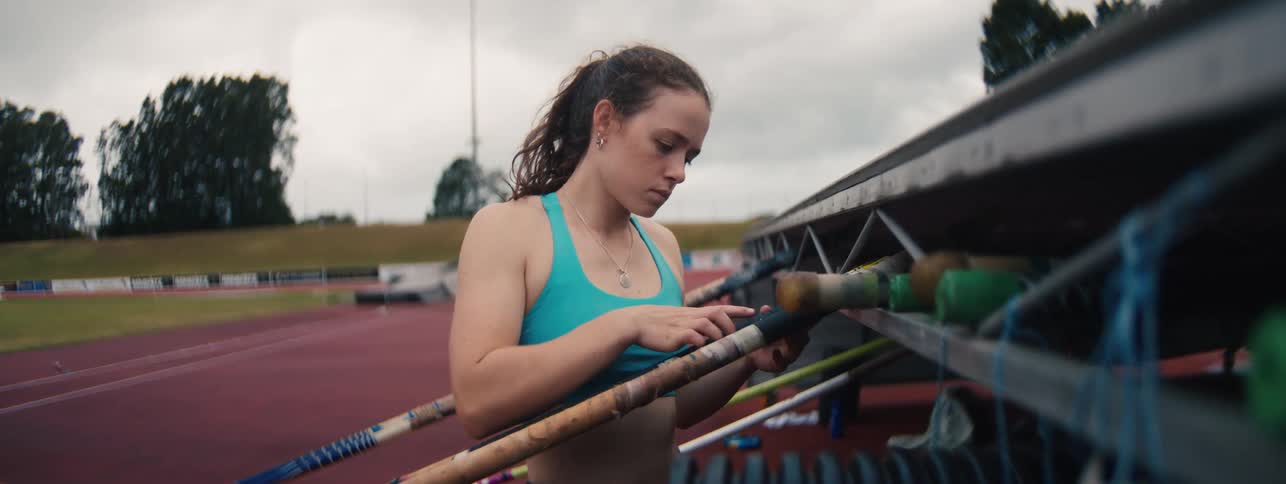 Female pole vaulter carefully examines and selects her pole from a rack in slow motion at an athletic stadium