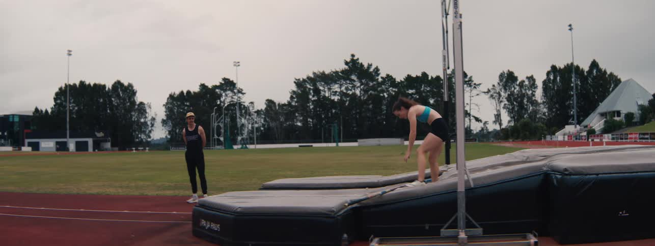 A male coach instructs his female athlete as she prepares for a pole vault attempt in slow motion at an athletic stadium
