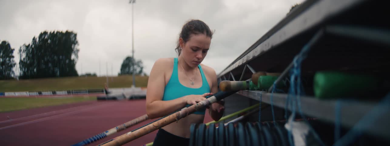 A determined female pole vaulter carefully selects her pole from a rack before training on an athletics track in slow motion